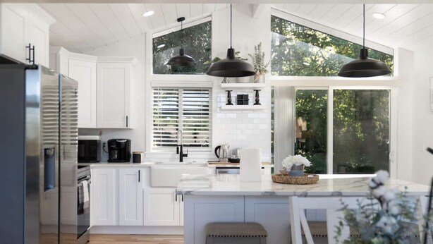 A modern kitchen with white cabinetry, a central island, pendant lighting, and large windows offering a view of greenery.
