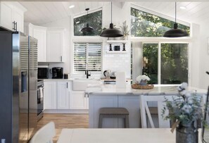 A modern kitchen with white cabinetry, a central island, pendant lighting, and large windows offering a view of greenery. - Delightful Family Home with 3 Bedrooms and Sprawling Backyard (Edmonds)