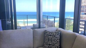 A modern living room with a beige sectional sofa and a patterned pillow, large windows offering a view of the ocean and a balcony with outdoor furniture. - Boardwalk Burleigh Beach 1 bed (Burleigh Heads)