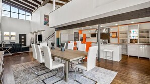 Un vaste espace de vie à aire ouverte comprenant un coin repas avec une table et des chaises blanches, ainsi qu’une cuisine équipée d’appareils électroménagers modernes.