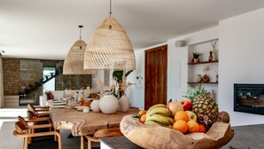 Una cocina moderna con un frutero de madera sobre la encimera, un comedor con sillas, y lámparas colgantes de mimbre.