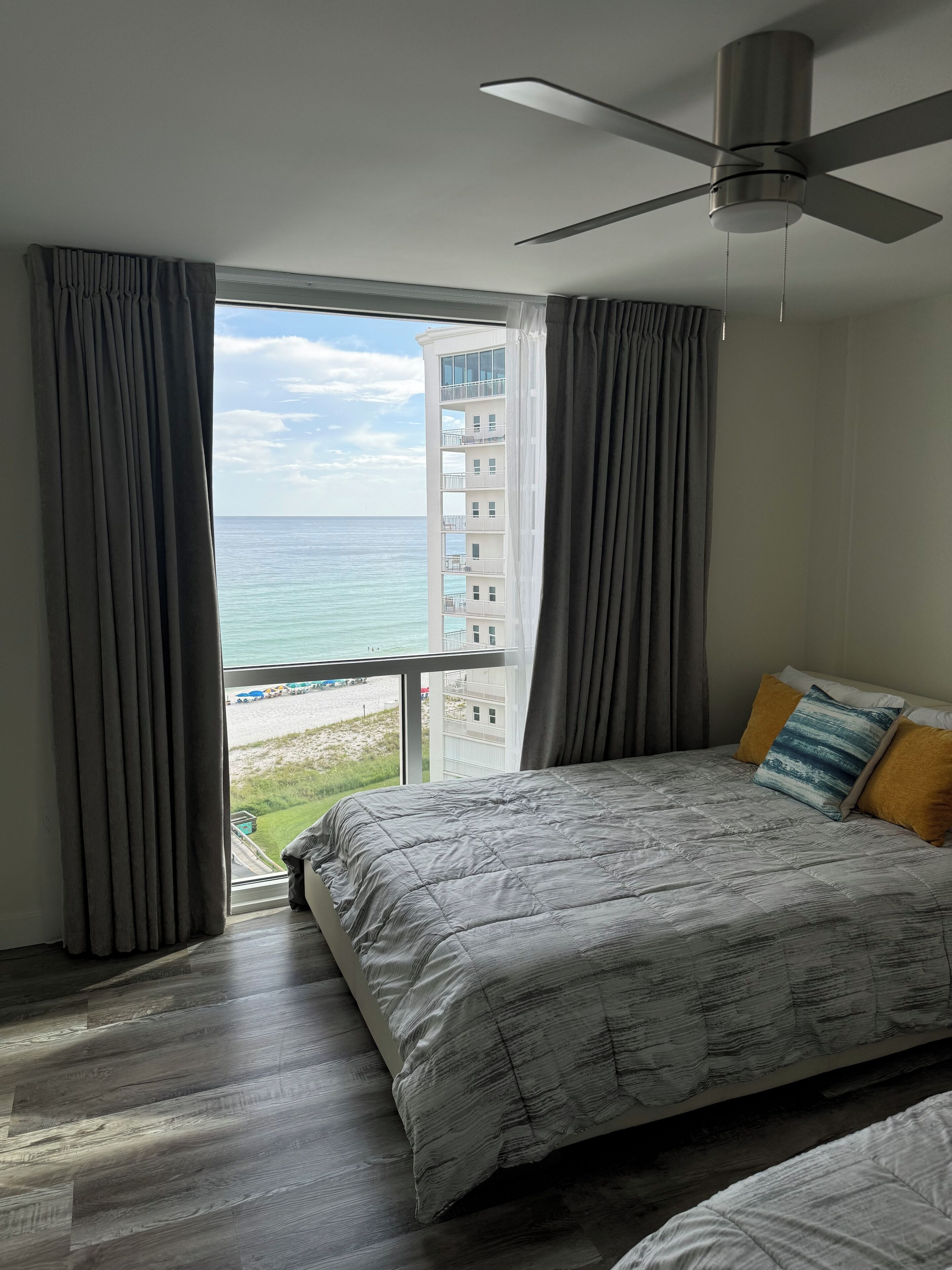 A bedroom with a bed, a ceiling fan, and a view of the ocean and buildings.