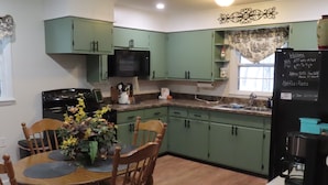 Una cocina con armarios verdes, una mesa de comedor con sillas, un televisor y un refrigerador con una pizarra.