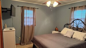 Un dormitorio con cama, mesita de noche, cómoda, ventilador de techo y ventana con cortinas.