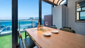 A dining area with a view of the beach and city skyline, featuring a wooden table, chairs, and a bottle of wine. - Goseong Badasga per Osyeonbyu Pension (Goseong)