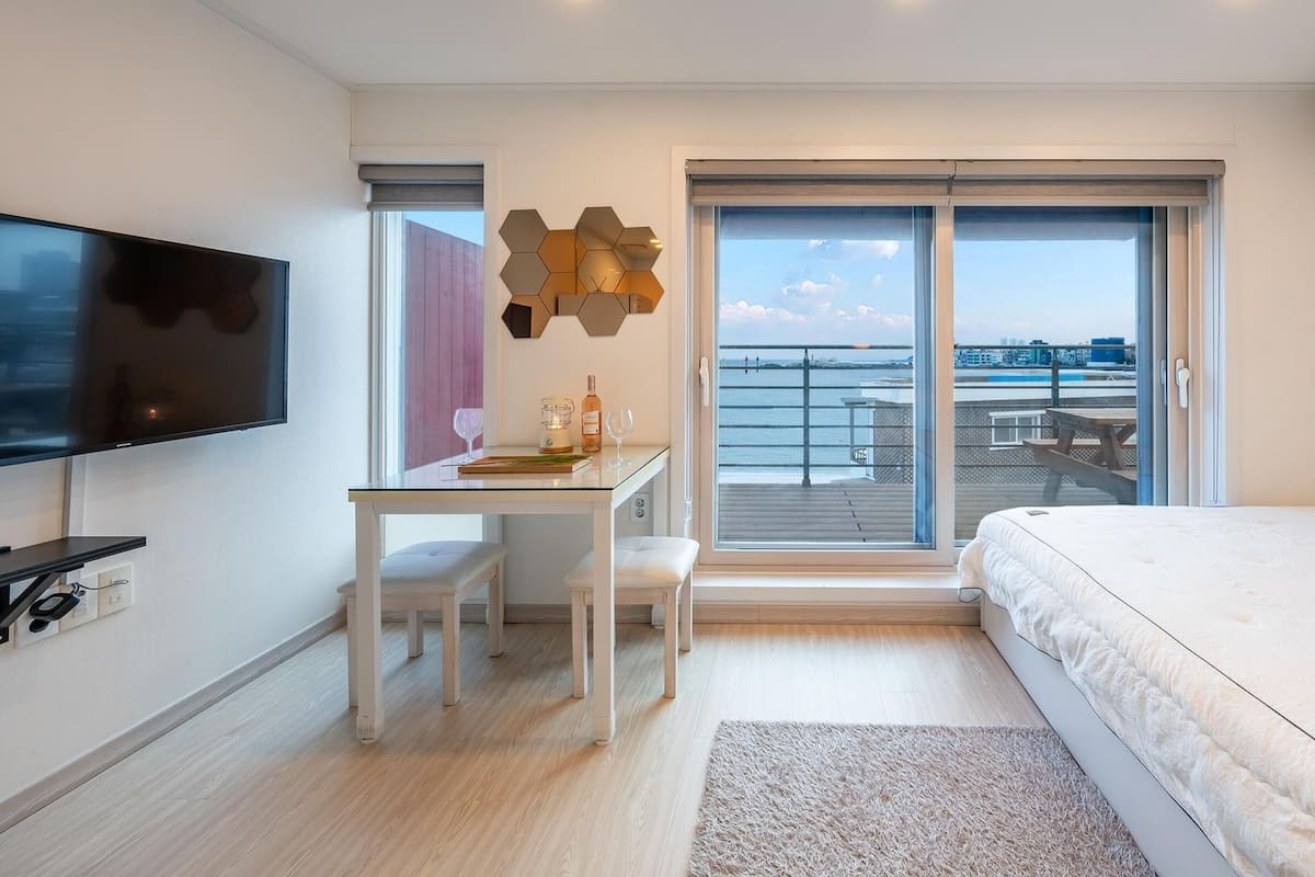 A modern hotel room with a flat-screen TV, a desk with a chair, a bed, and a large sliding glass door leading to a balcony with a view of the sea.