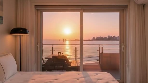 A hotel room with a bed, a nightstand, a lamp, a balcony with a table and chairs, and a view of the sunset over the water. - Goseong Badasga per Osyeonbyu Pension (Goseong)