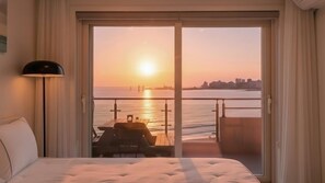 A hotel room with a bed, a nightstand, a lamp, a balcony with a table and chairs, and a view of the sunset over the water. - Goseong Badasga per Osyeonbyu Pension (Goseong)