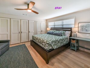 A bedroom with a bed, a ceiling fan, a desk with a lamp, and a window with blinds.