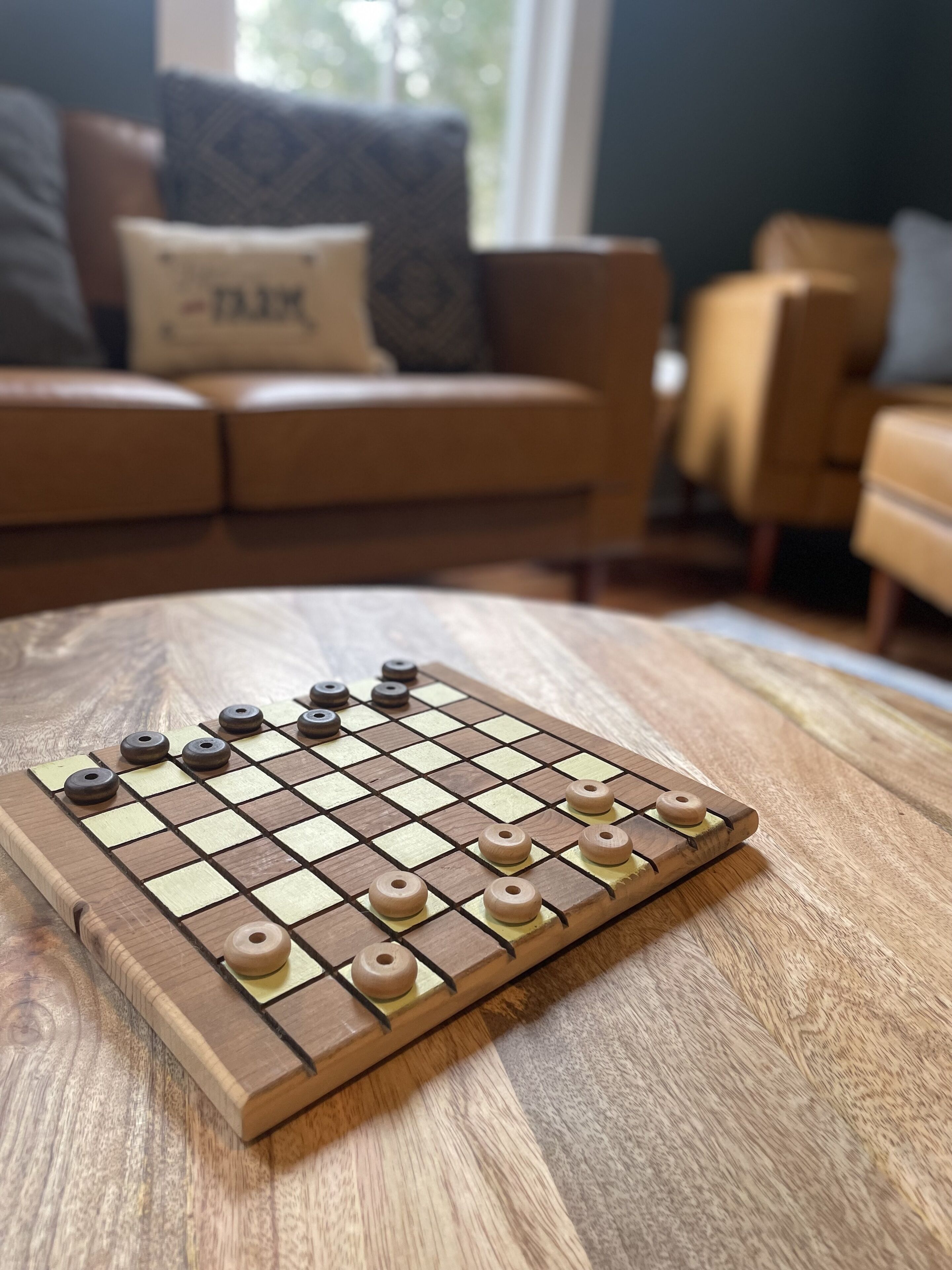 A wooden chessboard on a table in a living room with a sofa and a window in the background.