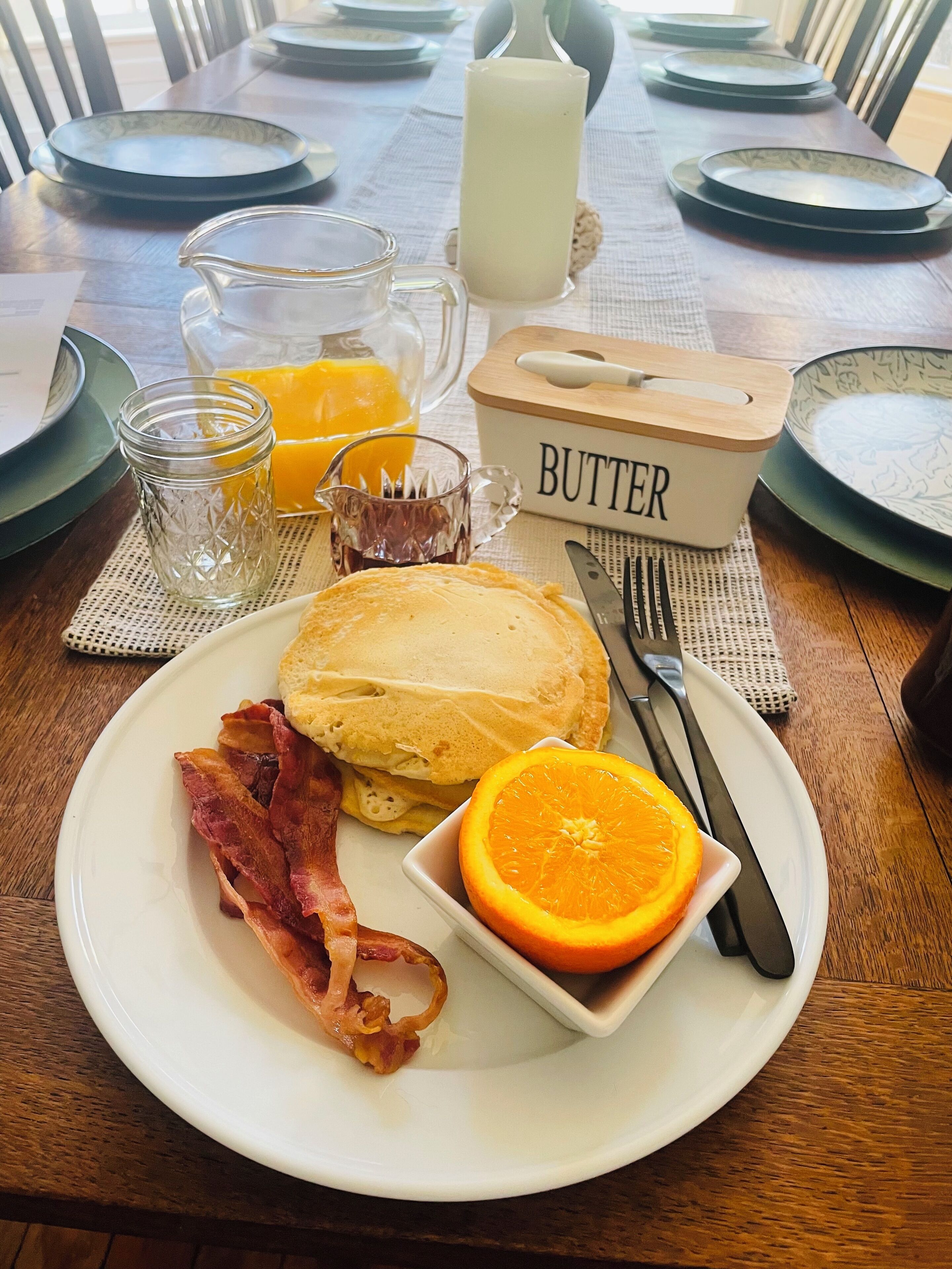 Una mesa de madera con panqueques, tocino, naranja y un recipiente de mantequilla.