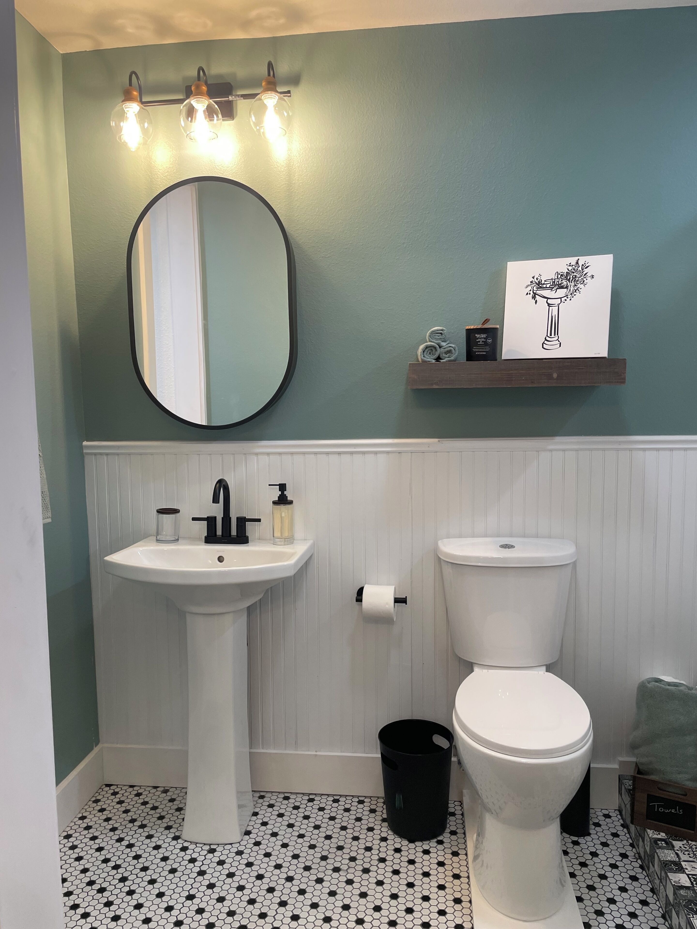 A bathroom with a pedestal sink, a toilet, a black trash bin, and a shelf with towels and a framed picture.