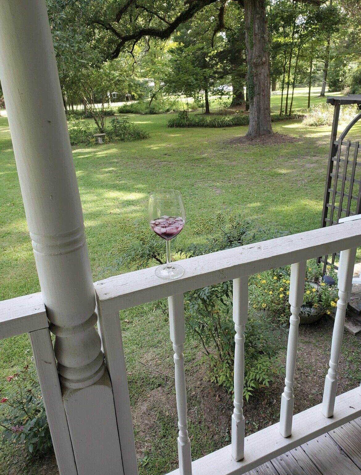 Um copo de vinho tinto sobre um corrimão branco, com vista para uma área gramada com árvores e um caminho.