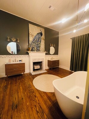 A bathroom with a freestanding bathtub, a round mirror, a peacock painting, a fireplace, and wooden flooring. - Adelines Bed and Breakfast (Coushatta)