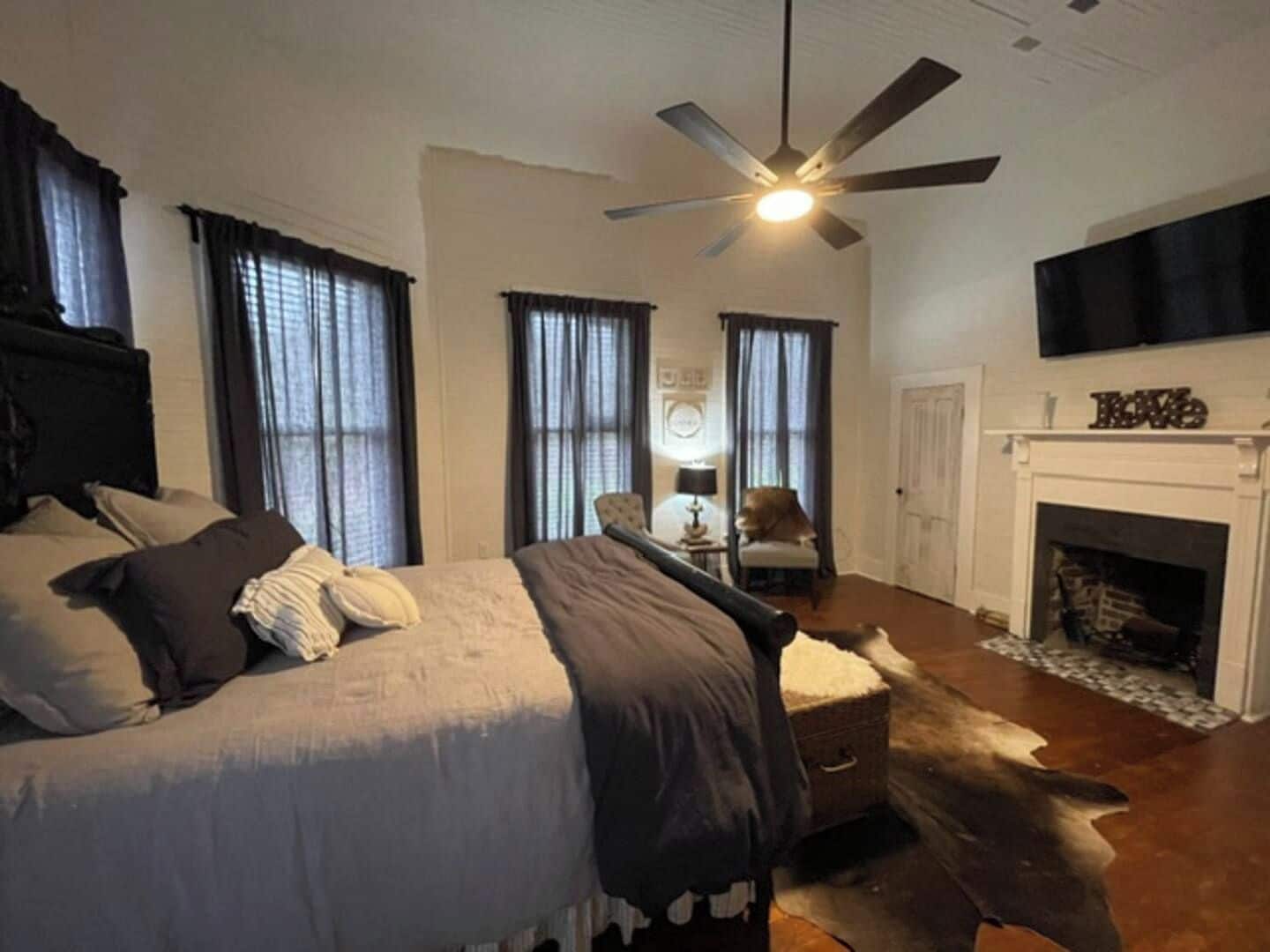 A bedroom with a bed, a ceiling fan, a fireplace, and a television mounted above it.