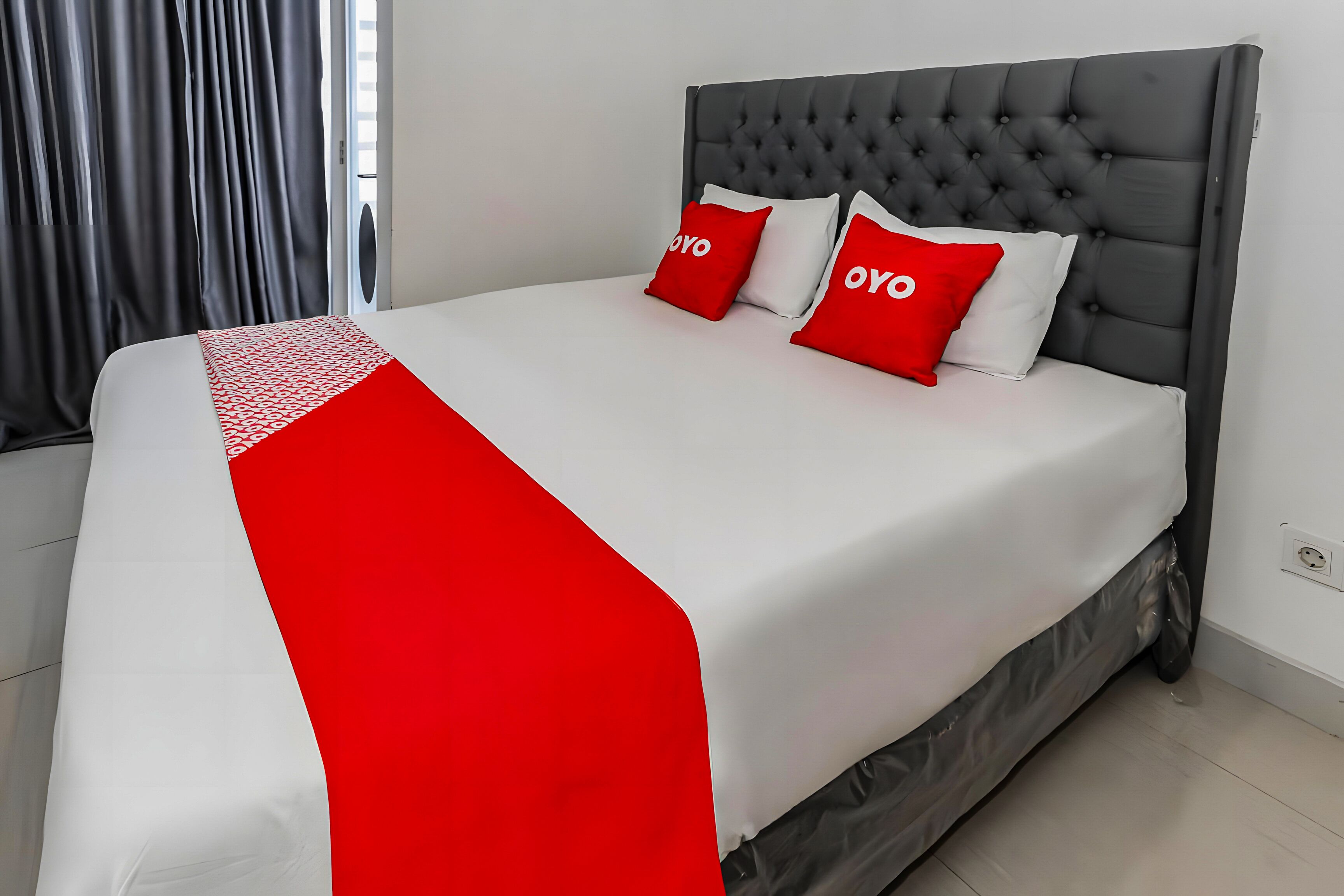 A neatly made bed with a grey upholstered headboard, white and red bedding, and two red pillows with the word "OYO" on them.