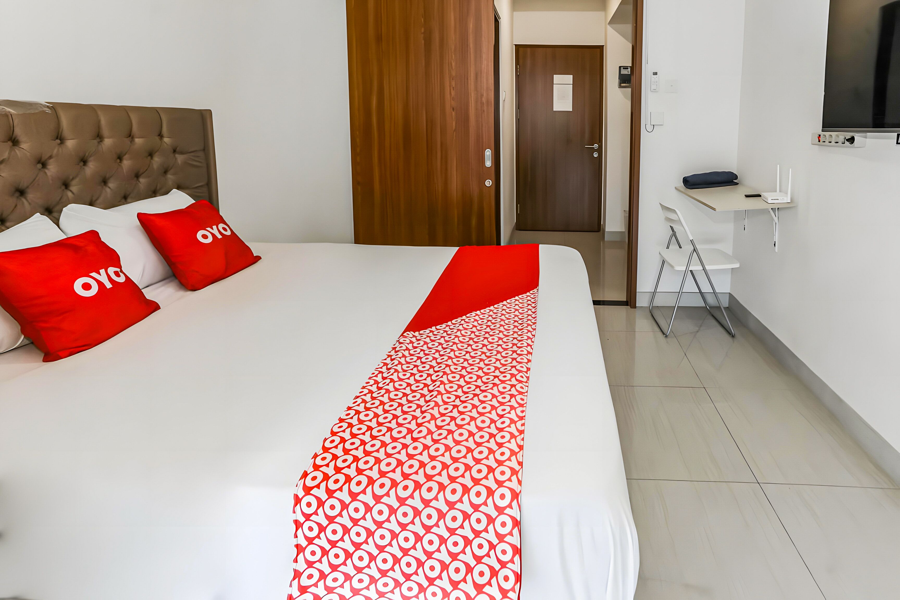 Una habitación de hotel ordenada con una cama grande, ropa de cama roja y blanca, una puerta de madera, un escritorio con una silla y un televisor montado en la pared.