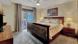 Un dormitorio con cama, ventilador de techo, mesita de noche, lámpara, televisor y vista al exterior.