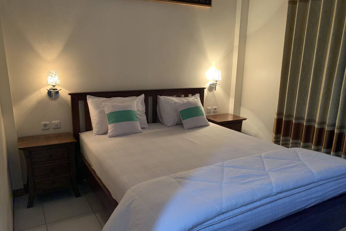 Una cama bien tendida con ropa blanca y dos almohadas, cabecera de madera, mesitas de noche con lámparas y una ventana con persianas.