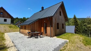 A wooden cabin with a sloped roof, a patio with a table and chairs, and a gravel driveway. - Pod lesem by Interhome (Smržovka)