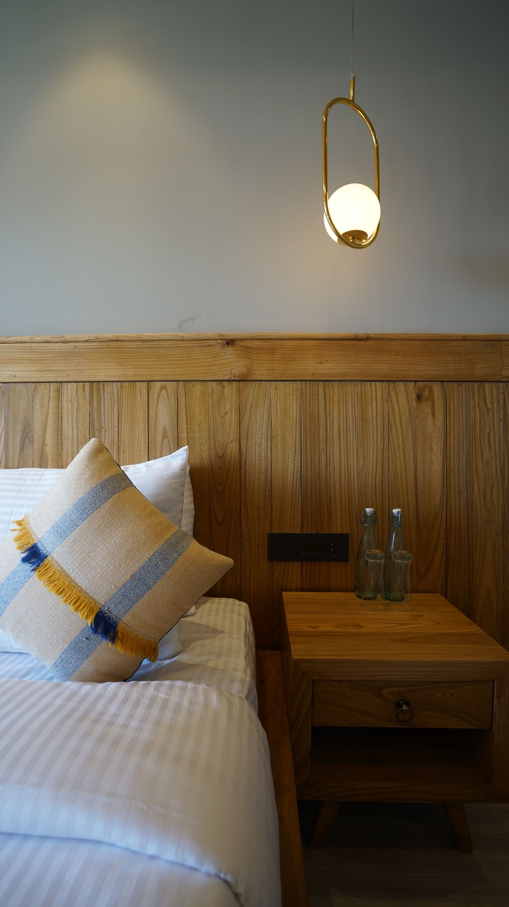 A neatly made bed with a patterned pillow, a wooden nightstand with bottles, and a wall-mounted light fixture.