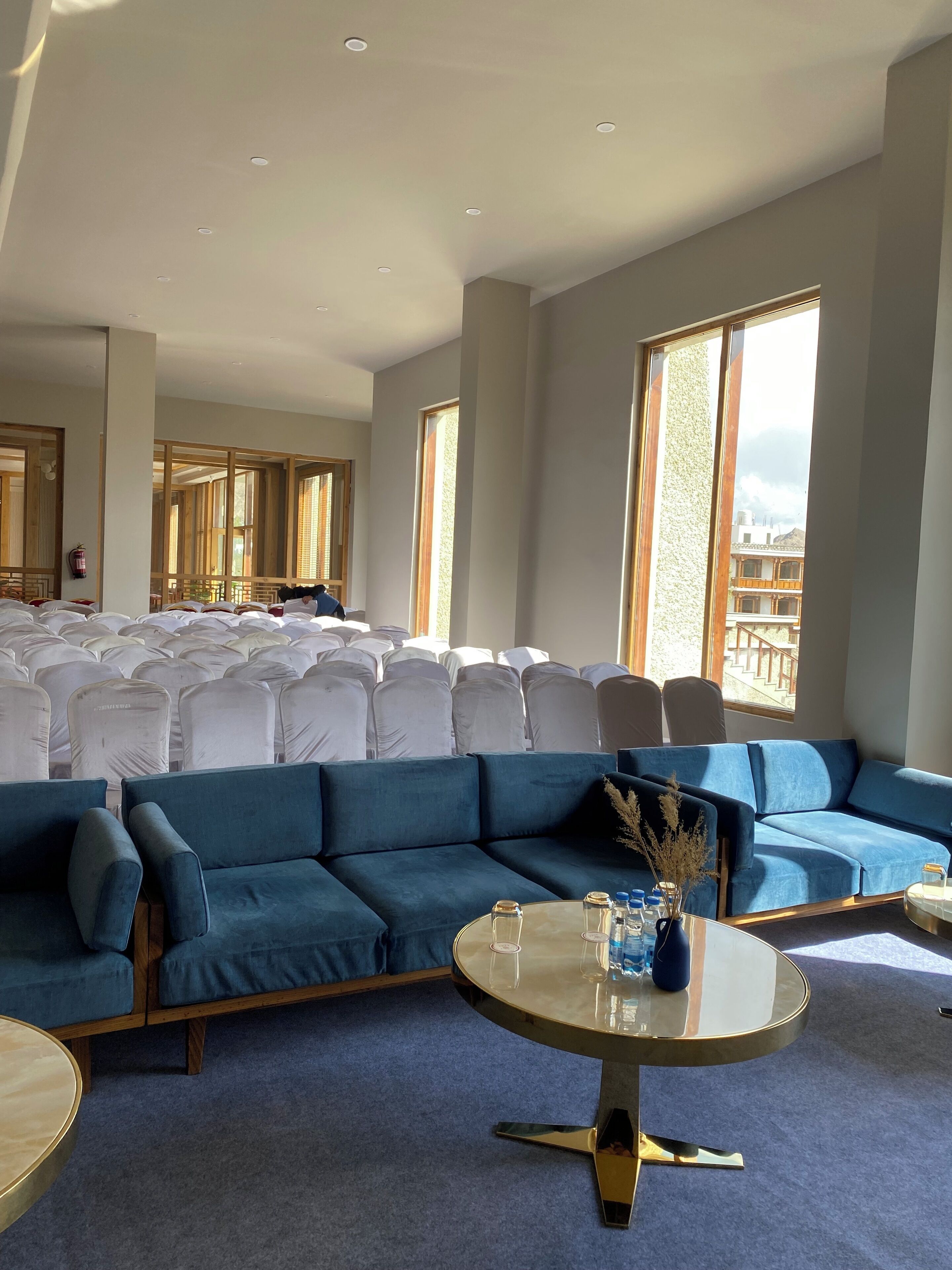 A spacious room with a large blue sofa, a round table with a vase, and a seating area with white-covered chairs.