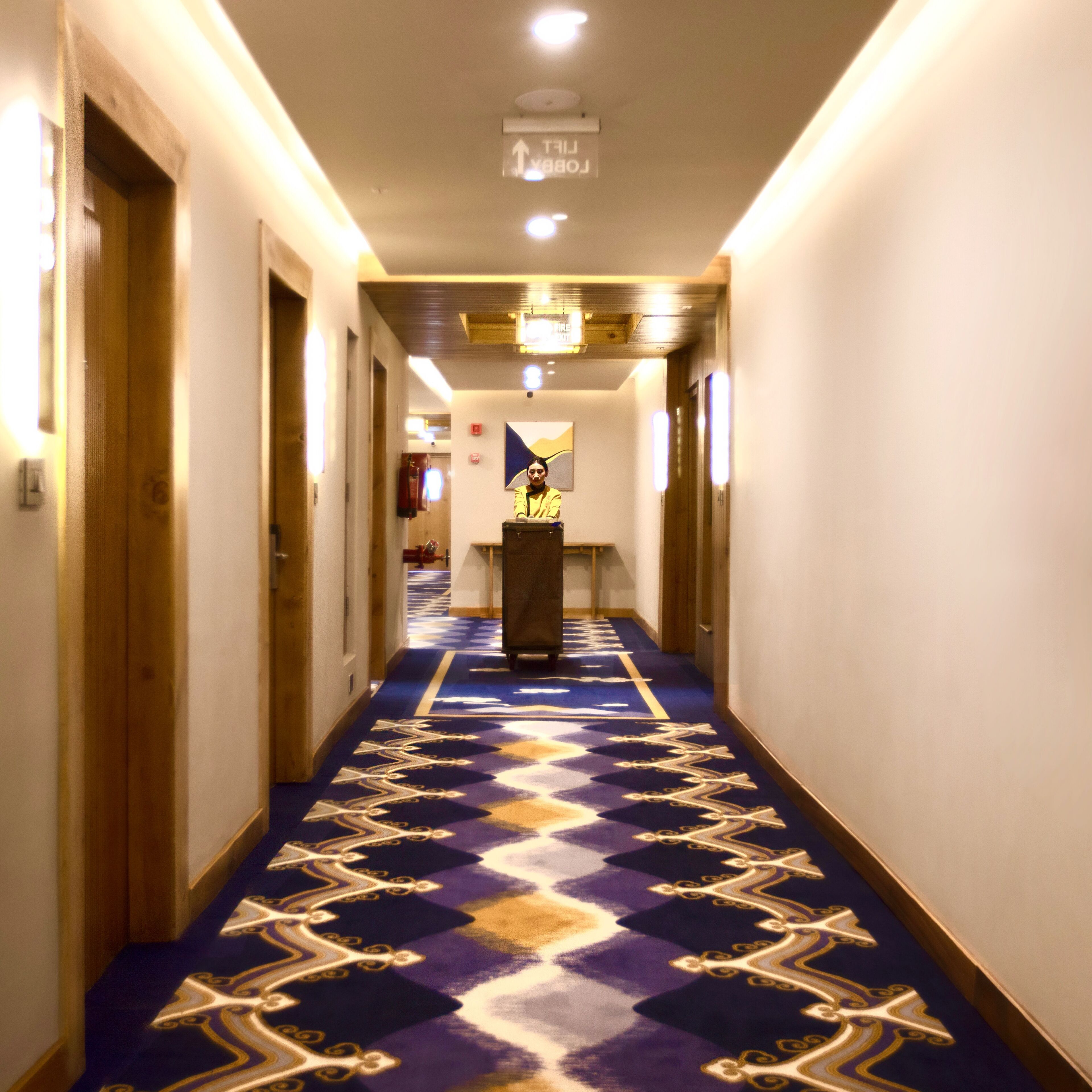 A hotel hallway with a patterned carpet, wooden doors, and a reception desk.