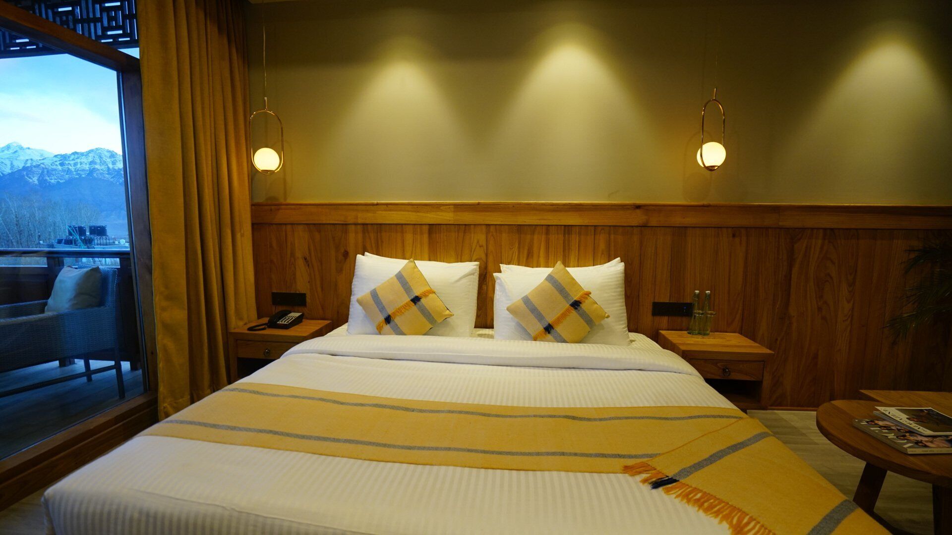 A hotel room with a large bed, two bedside tables, a nightstand, a window with curtains, and a view of mountains.