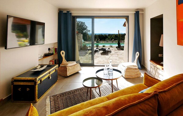 Un salon comprenant un canapé, une table basse, un téléviseur et donnant sur une piscine et un jardin grâce à une porte-fenêtre coulissante.