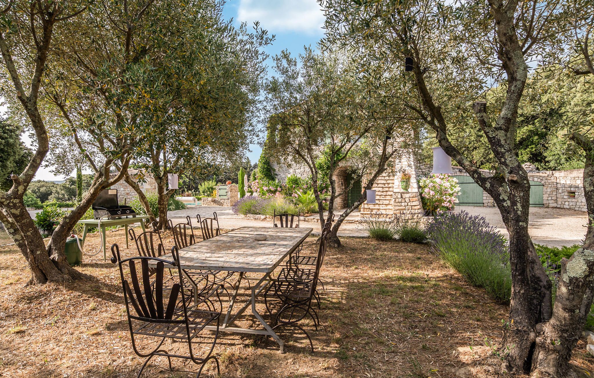 An outdoor dining area with a stone table and metal chairs, surrounded by trees and a stone wall.