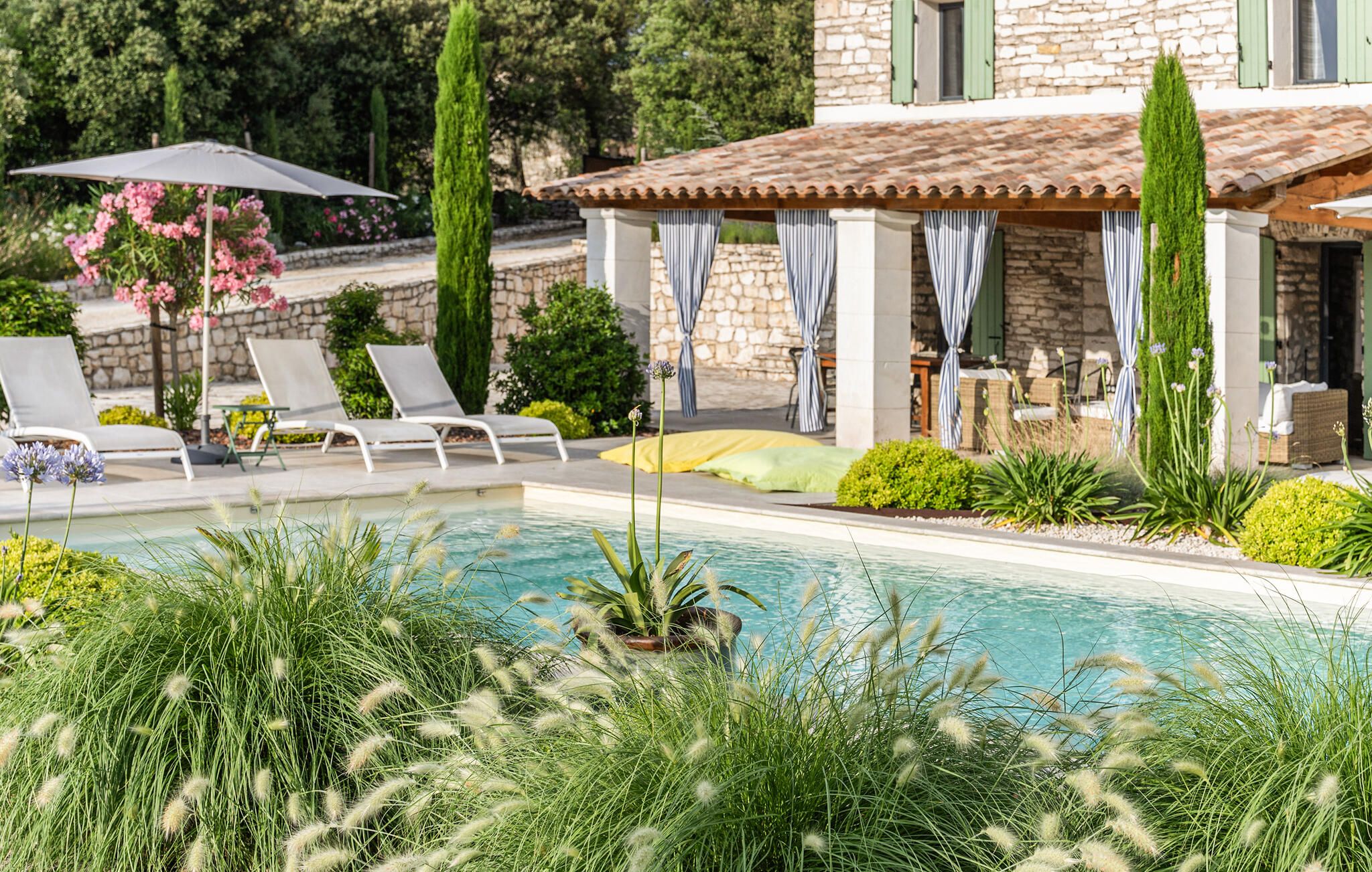 A pool area with lounge chairs, a covered seating area, and a stone building with a tiled roof.