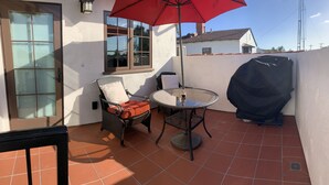 Un patio con una mesa de vidrio, dos sillas de mimbre, un paraguas rojo y una parrilla.