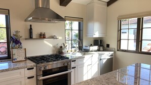Una cocina moderna con armarios blancos, una campana extractora de acero inoxidable, una estufa a gas, un horno y un amplio ventanal que proporciona luz natural.