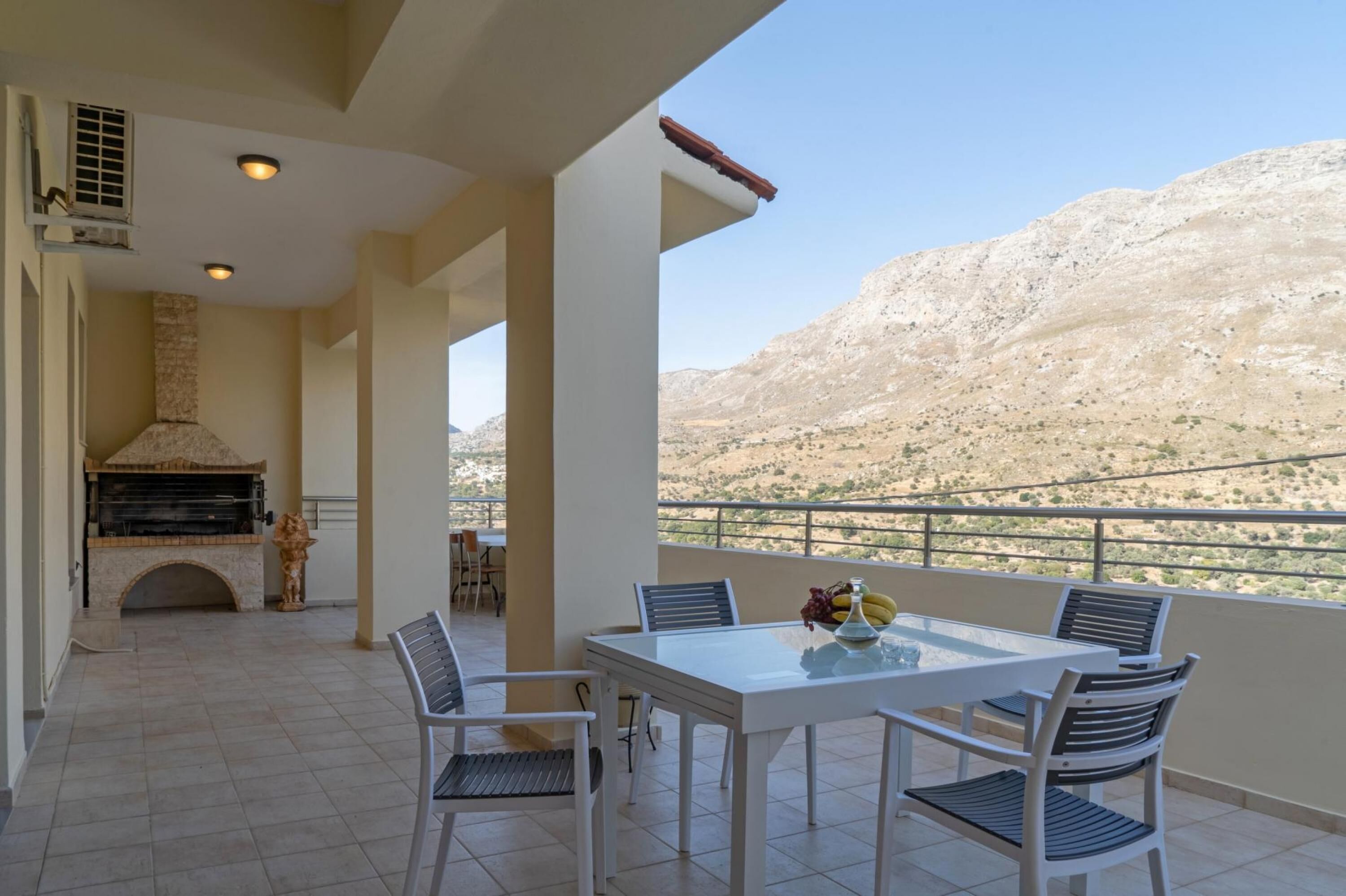 A balcony with a table and chairs, a grill, and a view of a mountainous landscape.