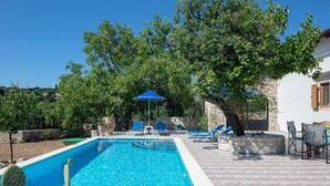 A swimming pool with lounge chairs and a table, surrounded by trees and a stone wall. - Silver Gem Villa With Private Pool (Rethymno)