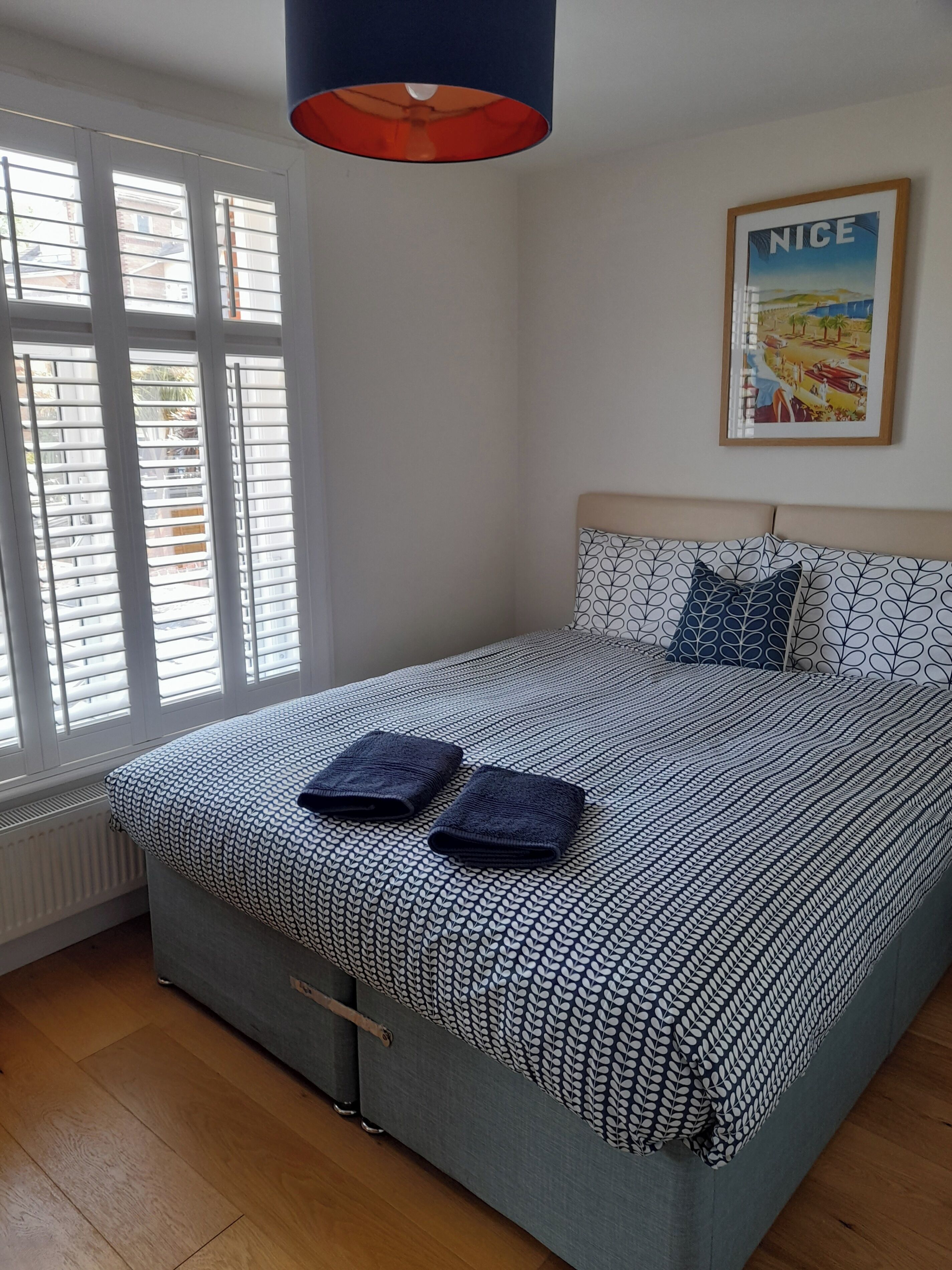Ein Schlafzimmer mit Bett, einem Fenster mit Jalousien, einem Heizkörper, einem gerahmten Bild an der Wand und einer Deckenleuchte.