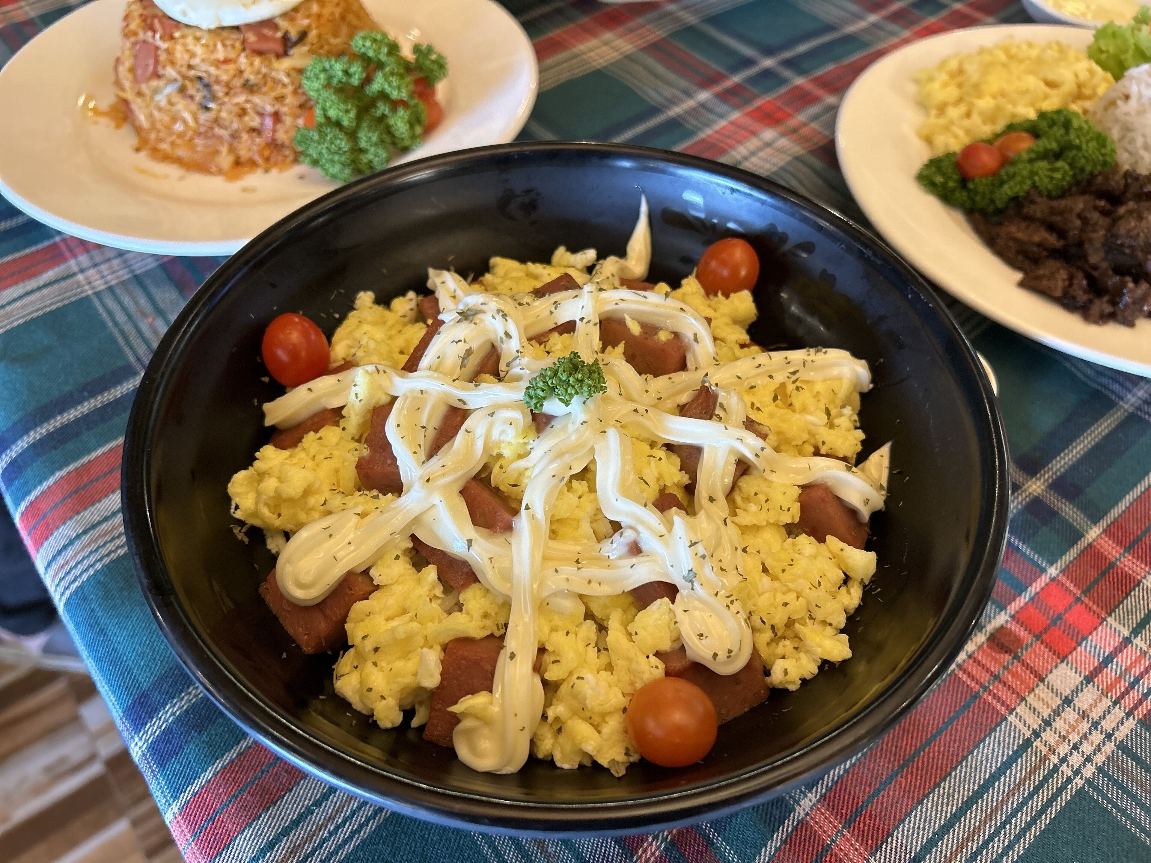A table with a plaid tablecloth, a black bowl of scrambled eggs with sausage and tomatoes, and other dishes in the background.