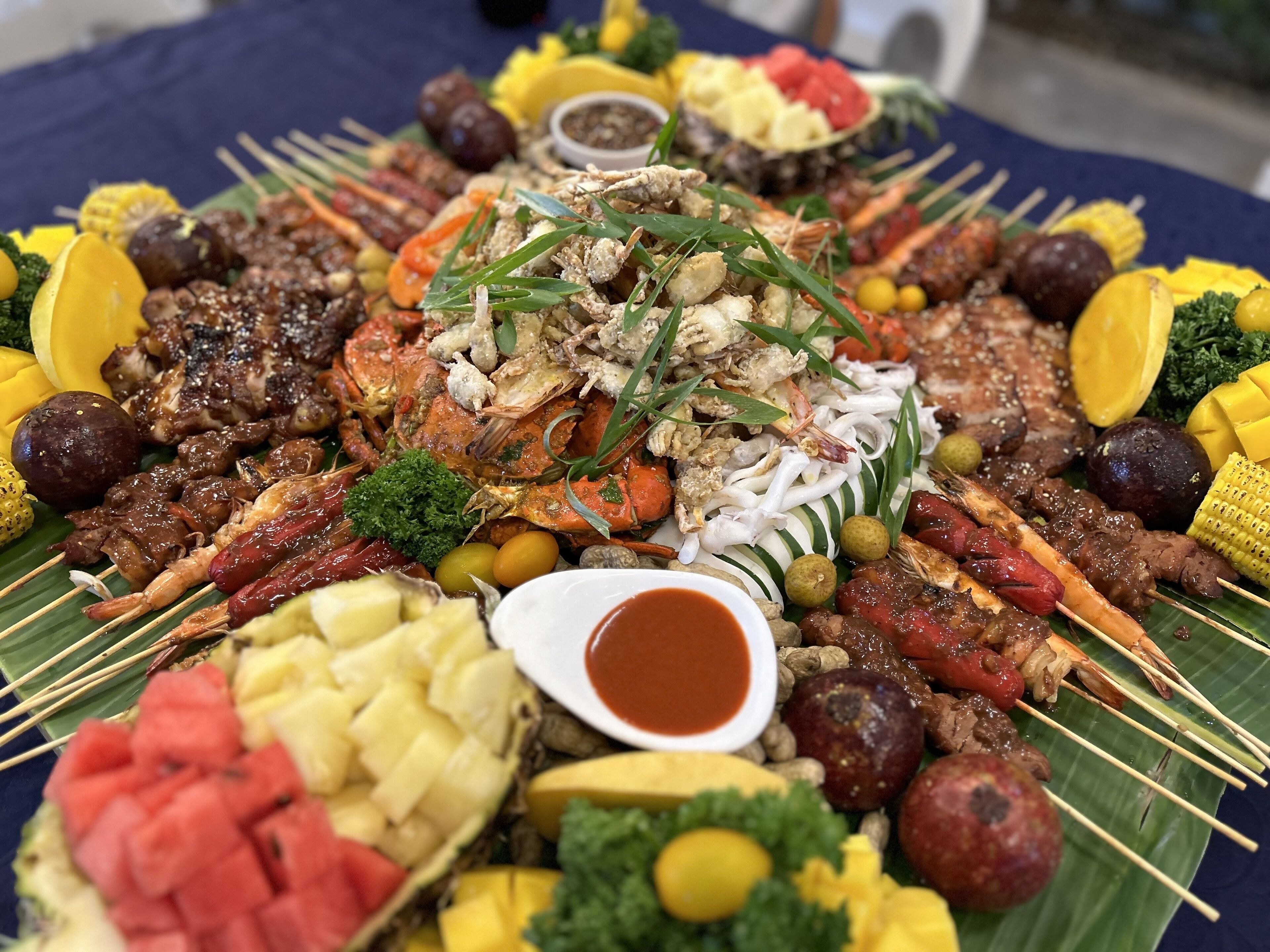 A large platter of various grilled meats, seafood, and vegetables, including corn, pineapple, and shrimp, arranged on a table.