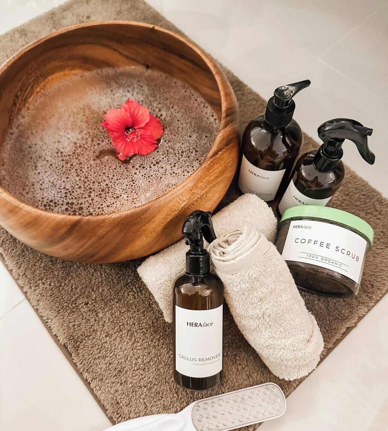 A wooden bowl with a red flower, a callus remover bottle, a coffee scrub container, a pump bottle, and a foot scrubger on a textured mat.