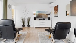 Un salon moderne avec deux fauteuils en cuir noir, un meuble de rangement blanc pour appareils multimédias, un téléviseur à écran plat et une grande fenêtre avec des rideaux.