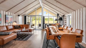 A spacious living area with a high ceiling, wooden walls, a large dining table, leather sofas, and a view through glass doors to the outdoors.