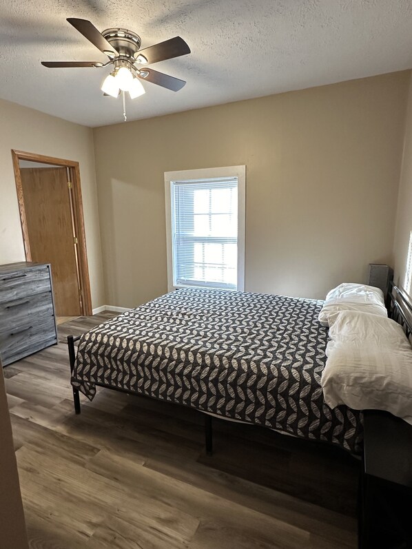 Un dormitorio con cama, ventilador de techo, ventana con persianas y puerta de madera.