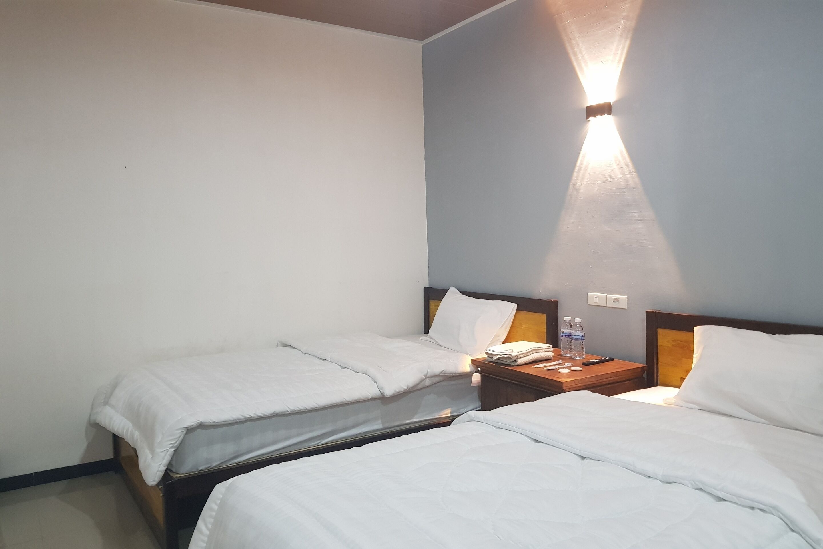 A hotel room with two single beds, a nightstand with a lamp, and a wall-mounted light fixture.