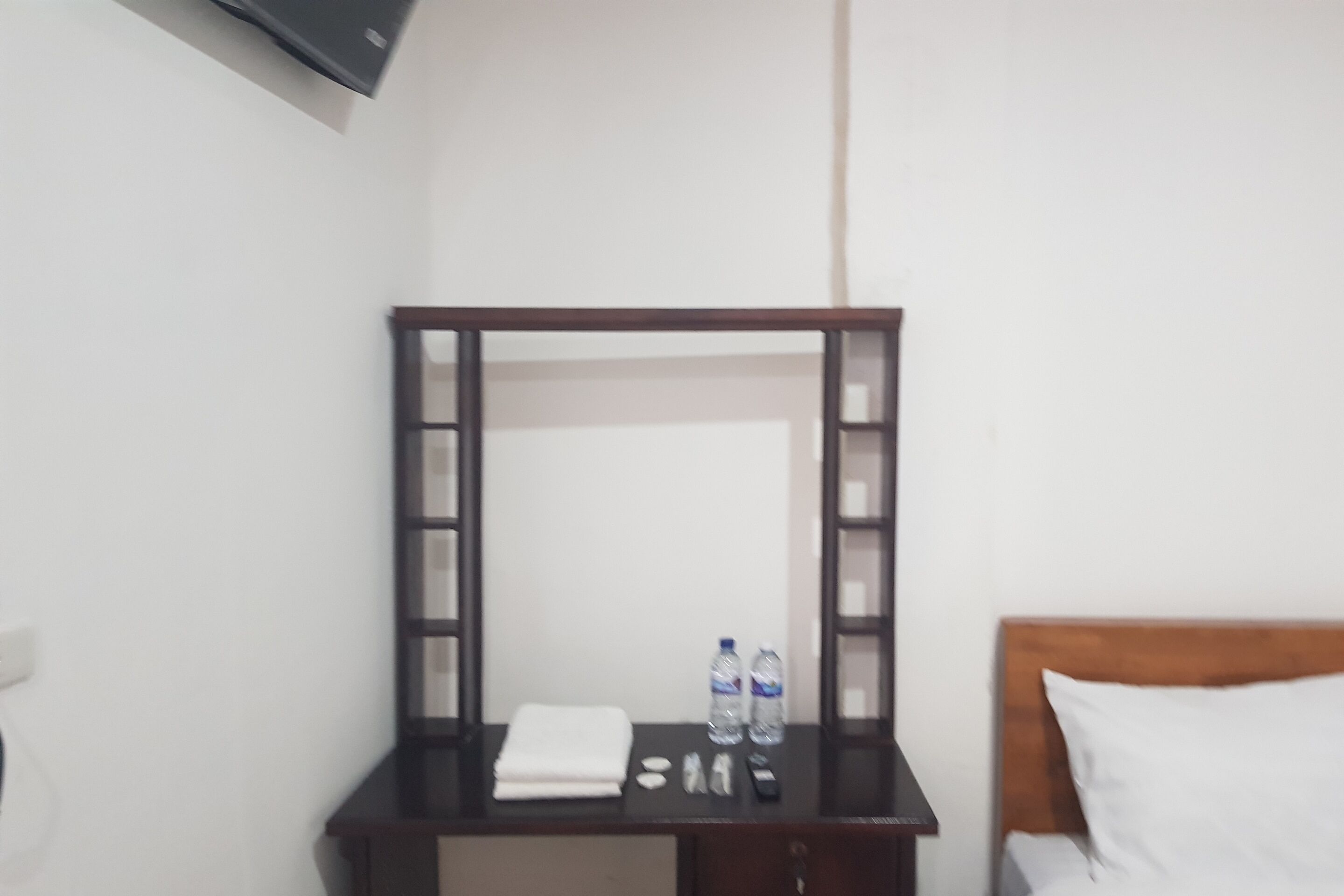 Ein Hotelzimmer mit einem Holztisch, einem Spiegel, einem Bett mit weißer Bettwäsche und einem Nachttisch mit Pflegeprodukten.