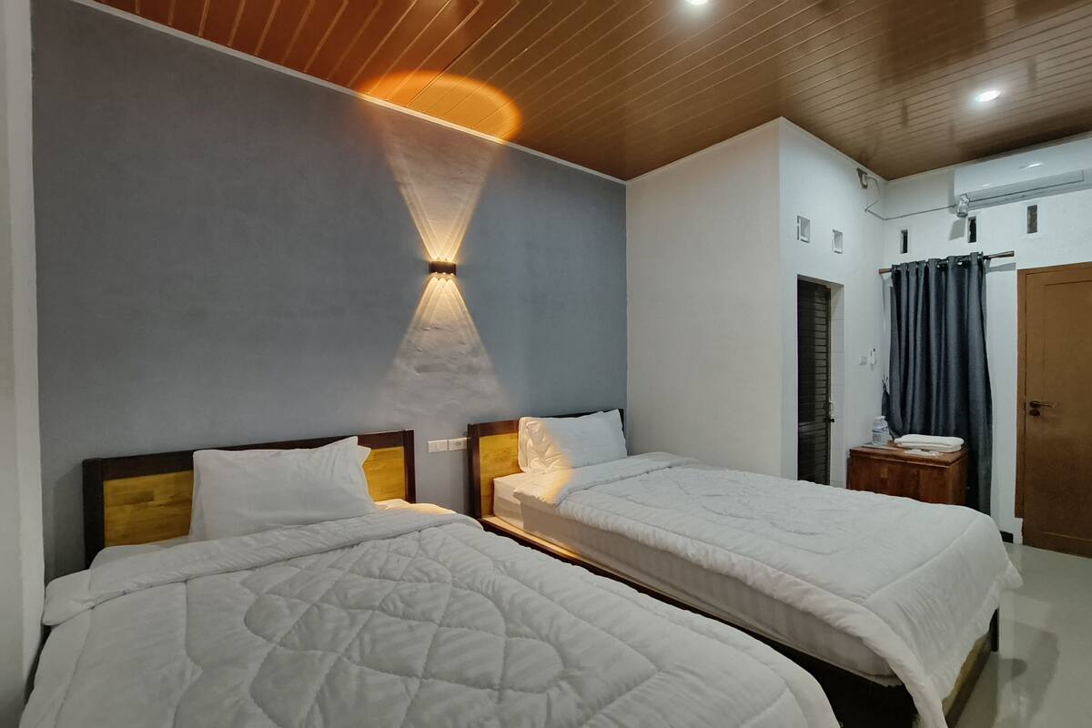 Une chambre d’hôtel avec deux lits, un plafond en bois, un luminaire fixé au mur, une petite table de chevet en bois, une porte et un rideau.
