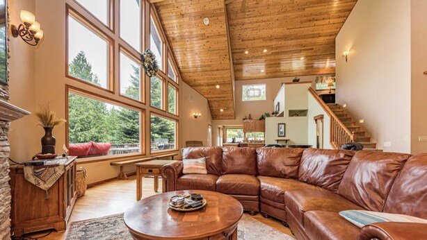 A spacious living room with a large leather sectional sofa, a wooden coffee table, and a fireplace. Large windows provide natural light and a view of the outdoors. - Majestic Sequim Vacation Home near Sequim Bay (Sequim)