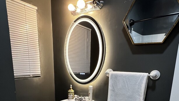 Un baño con lavamanos blanco, espejo con iluminación, toallero y pared oscura.