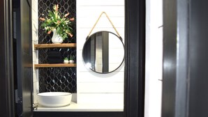 Un baño con lavamanos blanco, un espejo redondo y estantes con plantas.