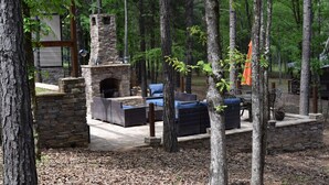 Una chimenea de piedra rodeada de asientos exteriores, ubicada en una zona boscosa con árboles y un muro de piedra.