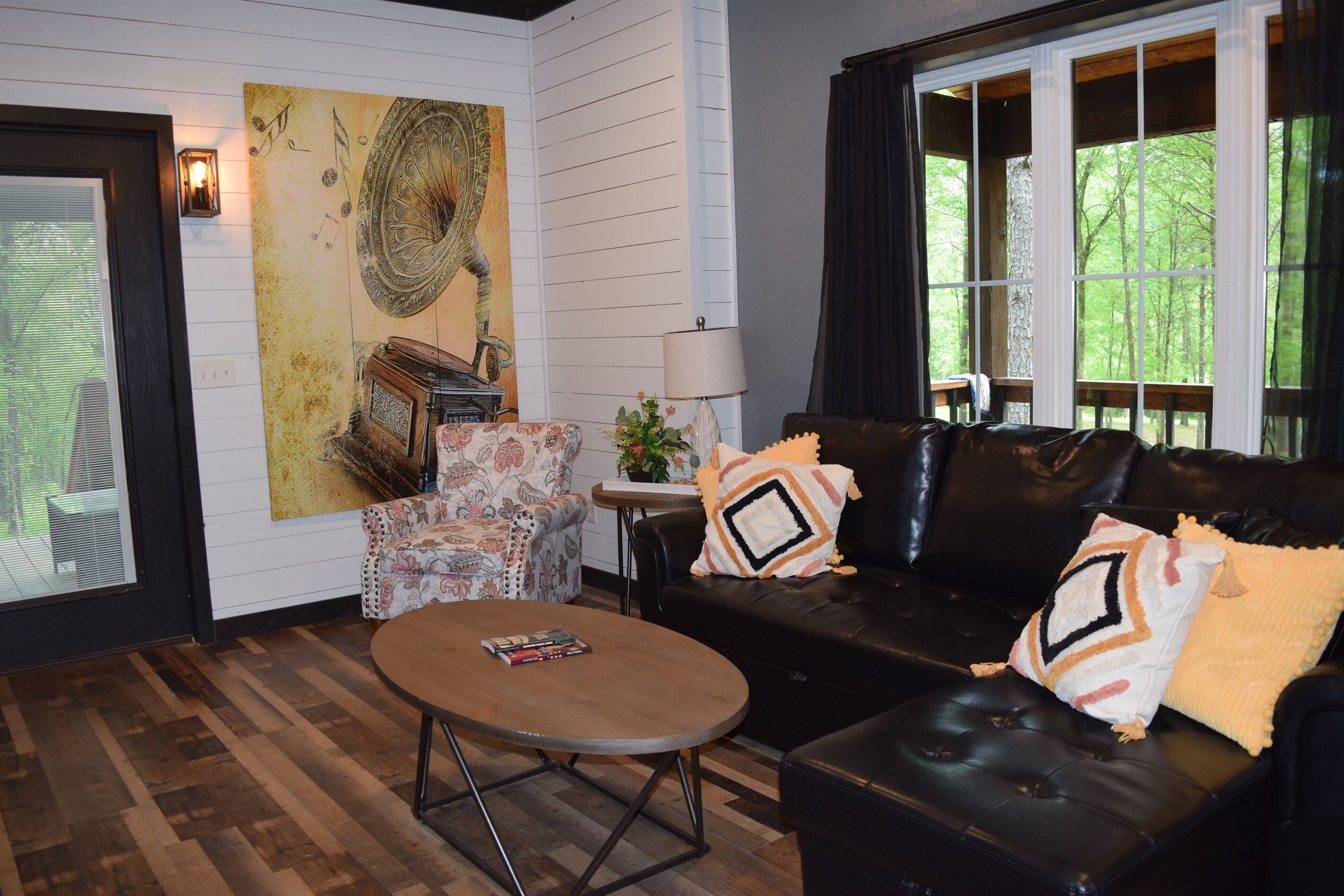 A living room with a wooden floor, a black leather sofa, a patterned armchair, a round coffee table, and a large abstract painting on the wall.